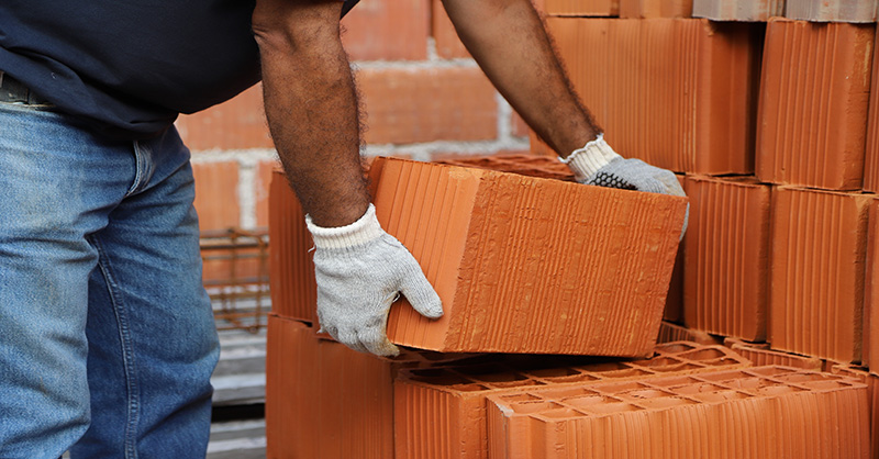 A person lifting a large brick with proper technique, showcasing the importance of ergonomics in manual labor. The individual is bending at the knees and keeping the back straight while wearing protective gloves