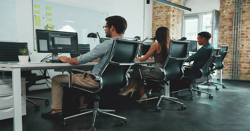 Three individuals seated on ergonomic chairs in front of desktop computers in a modern office setting.