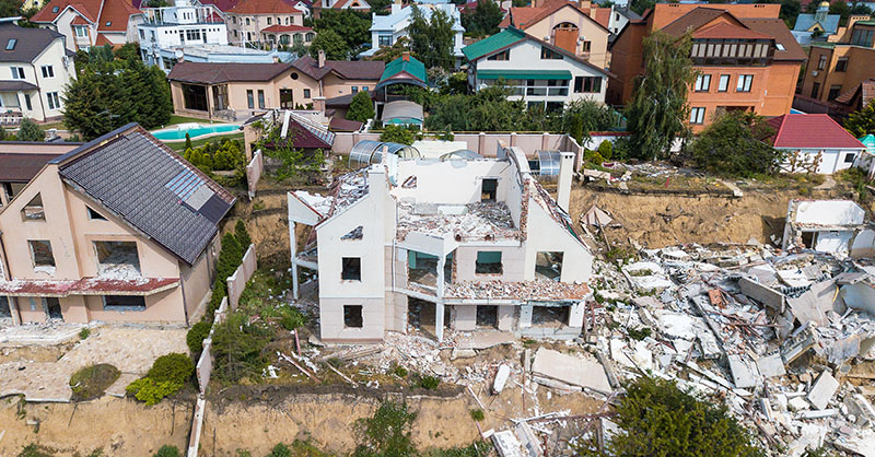 Aerial view of a residential area showing the aftermath of an earthquake with one house completely collapsed among others that have withstood the tremor, highlighting the importance of earthquake preparedness in building design and construction.