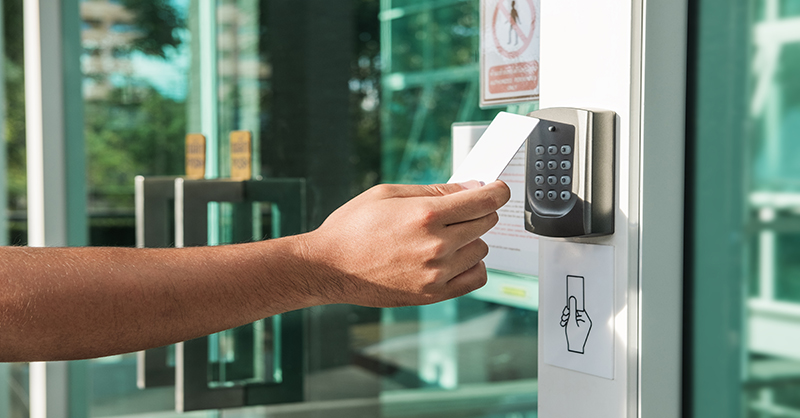 A person’s hand holding a white access card up to a black electronic card reader beside a glass door, symbolizing controlled entry as part of organizational security measures.