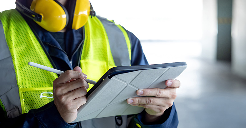 A person is holding a tablet and a stylus, presumably conducting a safety checklist. The individual is focused on the tablet screen, indicating attention to detail and adherence to safety protocols in an industrial or construction environment