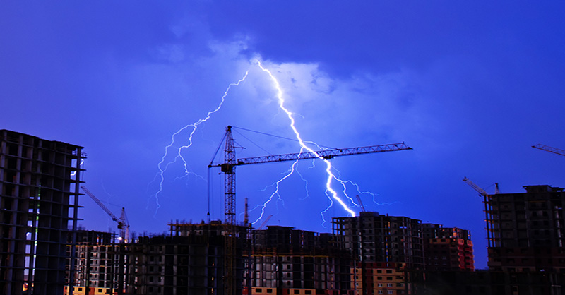 A construction site with multiple high-rise buildings and a vivid lightning bolt strikes down in the background, illuminating the scene and highlighting the potential dangers of severe weather at construction sites