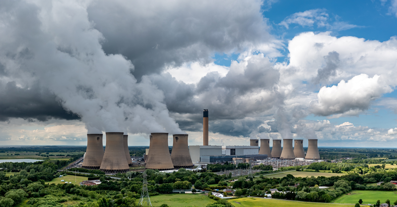 A panoramic view of a large power plant with multiple cooling towers emitting steam against a backdrop of a cloudy sky, illustrating the impact of industrial activity on the environment.