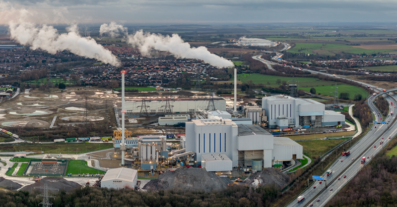 Aerial view of an industrial complex with multiple buildings, smokestacks emitting white smoke, a large pile of raw materials, and surrounding infrastructure including a highway.