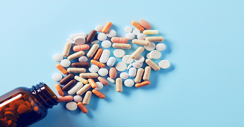 An assortment of various pills and capsules spilled from an open brown prescription bottle onto a light blue background. The medication forms a scattered pile, showcasing a range of sizes, shapes, and colors such as white, orange, and beige.